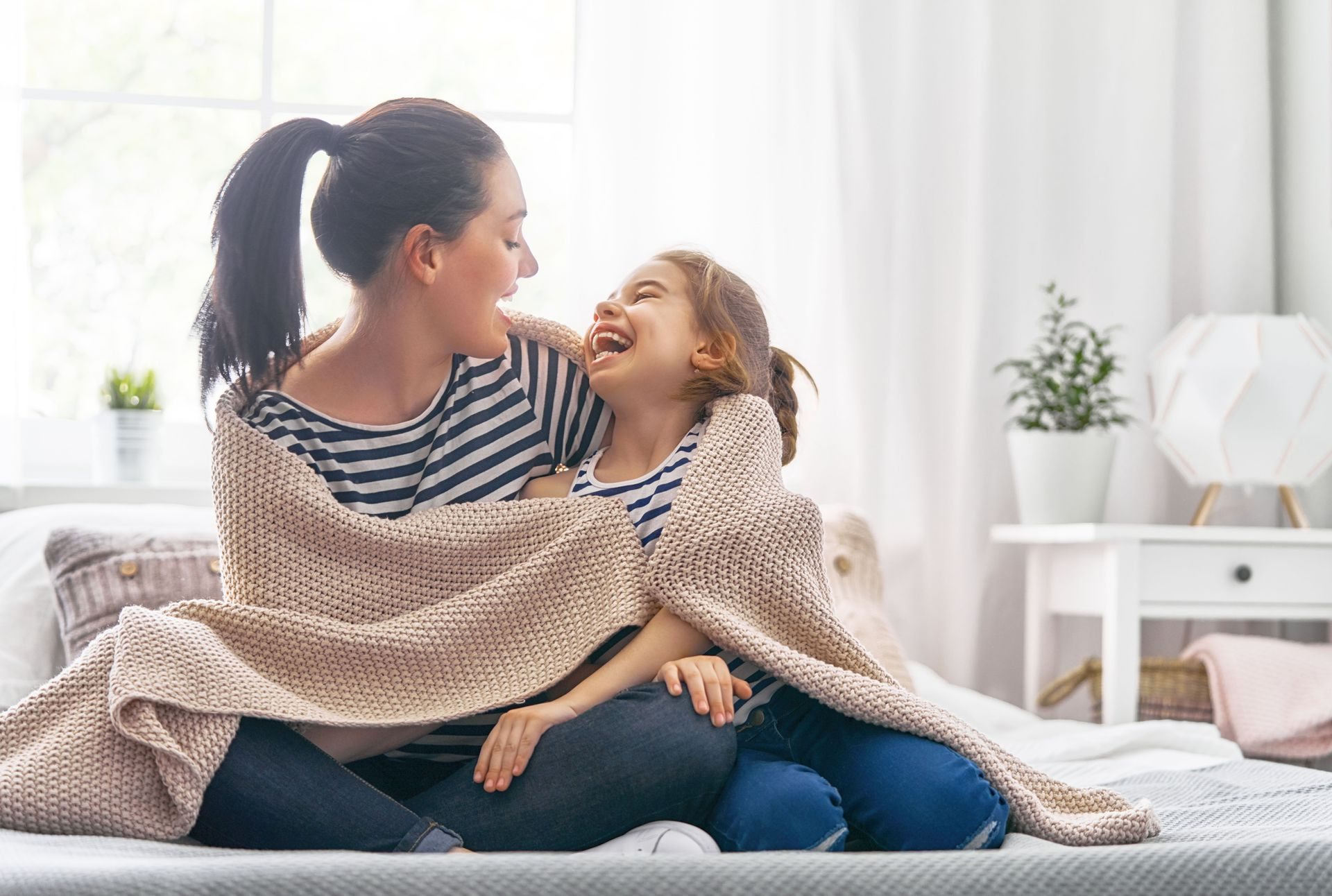 Woman and child laughing, wrapped in a blanket on a bed, sunny room.