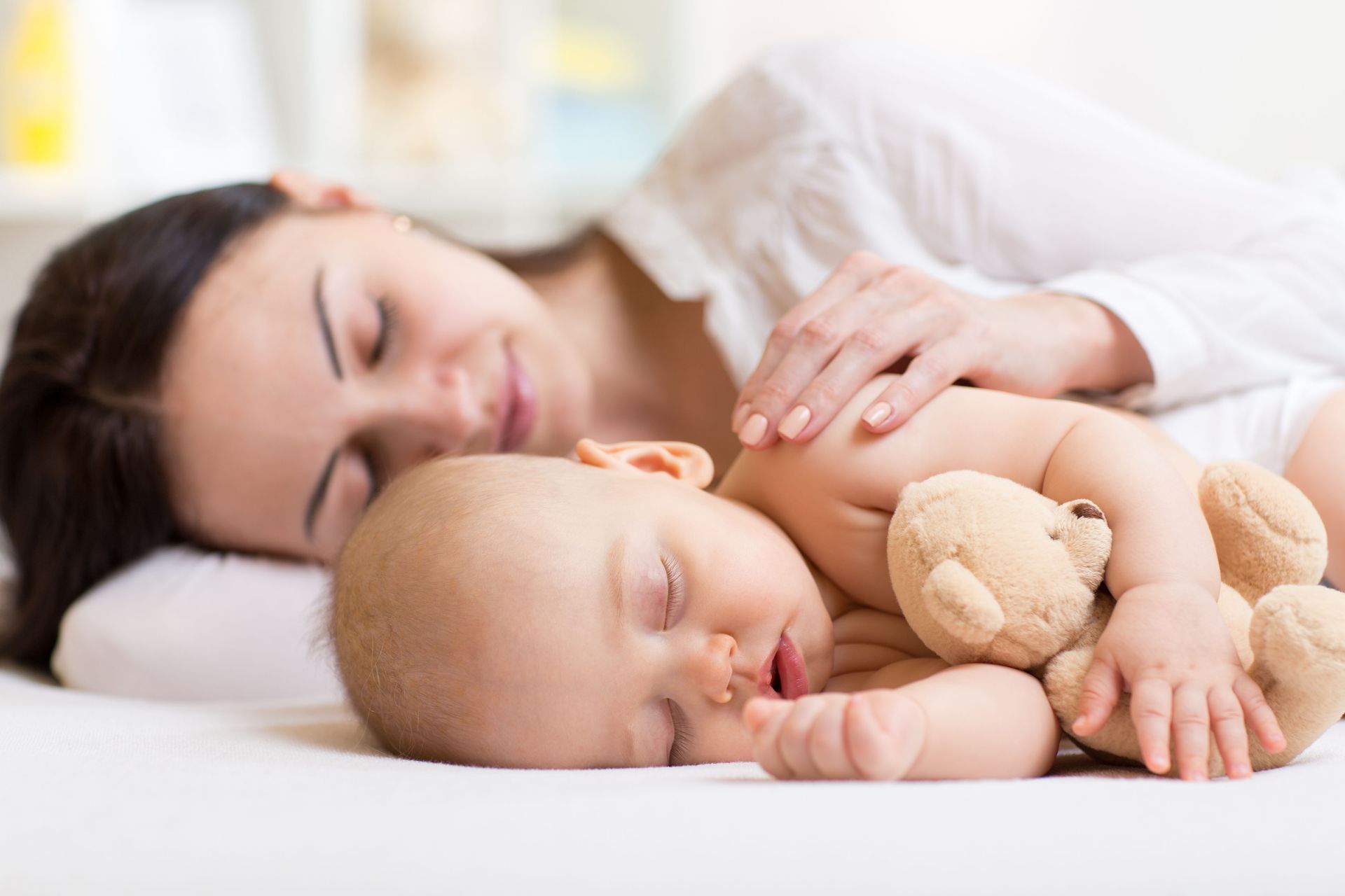 Mother and baby sleeping on a bed, baby holding a teddy bear, white bedding.