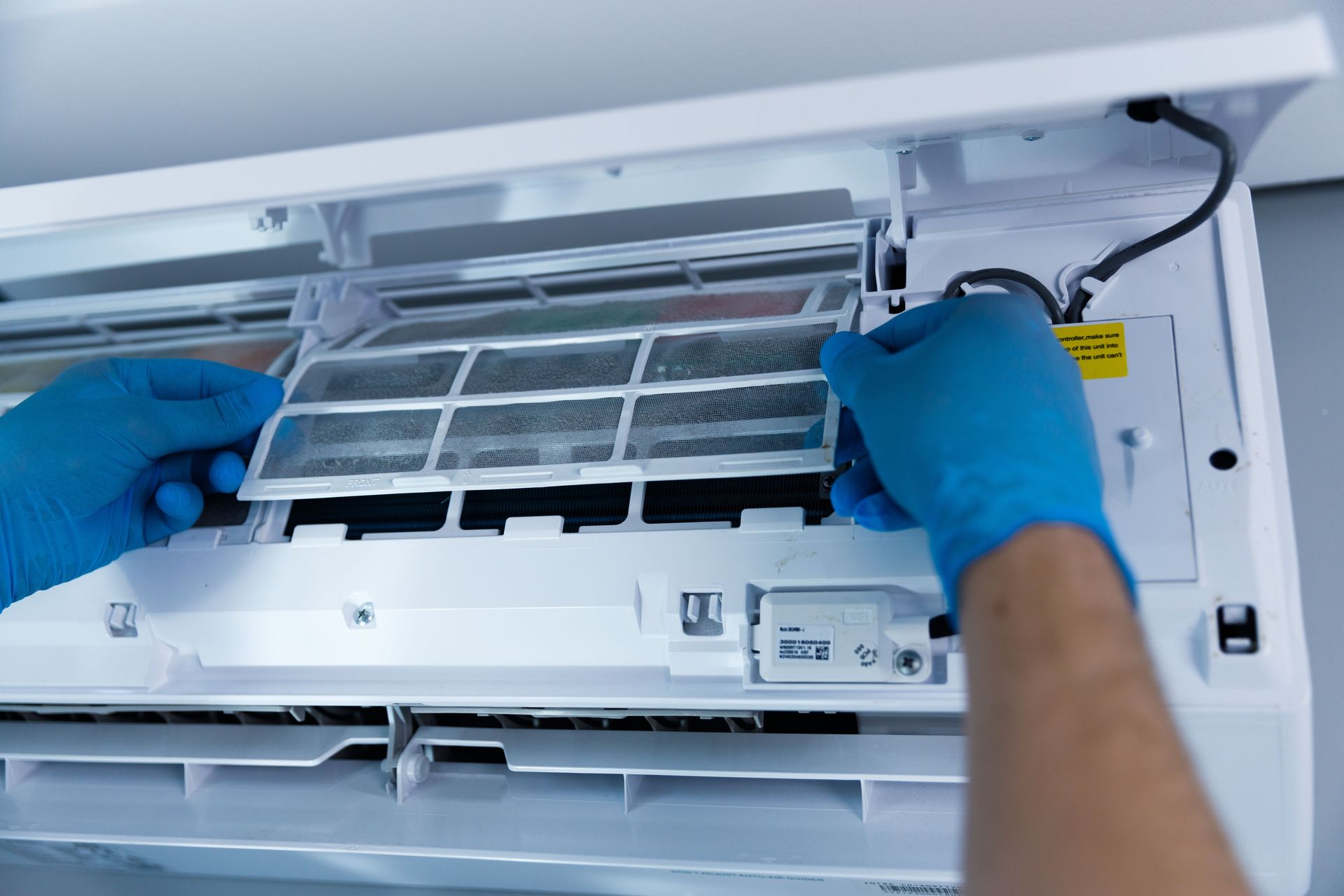 Hands cleaning an air conditioner filter inside the open unit with blue gloves