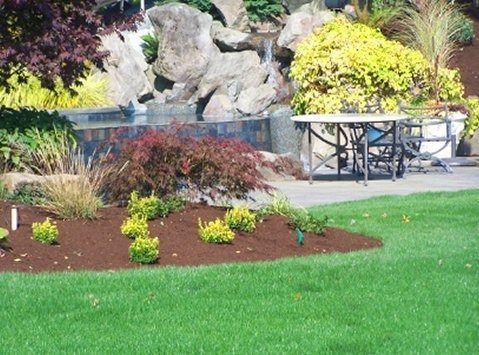 Green lawn, landscaped garden with waterfall, table and chairs. Lush greenery with red and yellow plants.