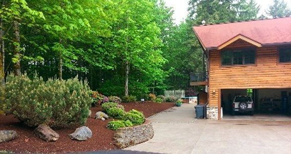 Wooden house with garage, lush greenery, and landscaped yard.