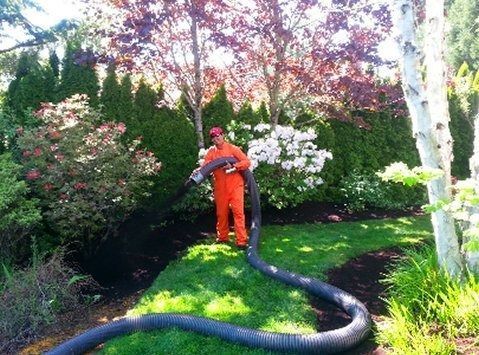 Person in orange jumpsuit with hose in garden bed; trees and shrubs in the background.