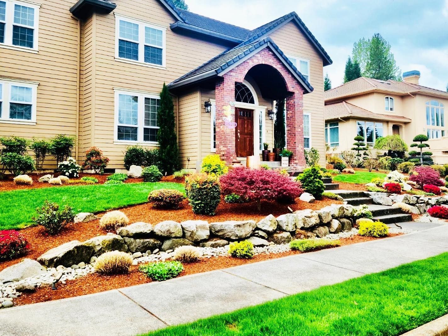 Two-story house with manicured lawn and colorful landscaping, including shrubs and a stone retaining wall.