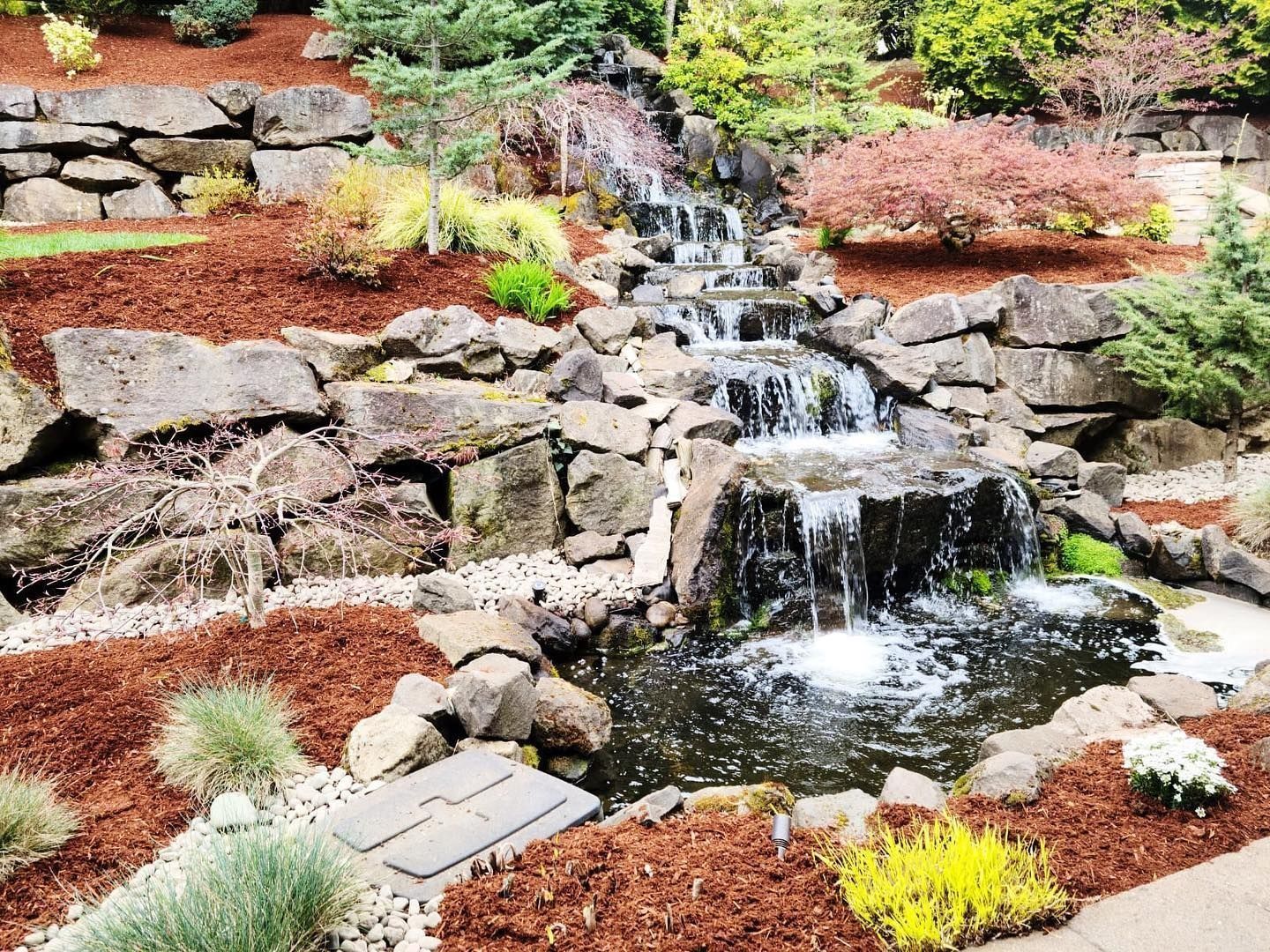 Stone waterfall cascading into a pond, surrounded by rocks and brown mulch, with greenery and colorful plants.