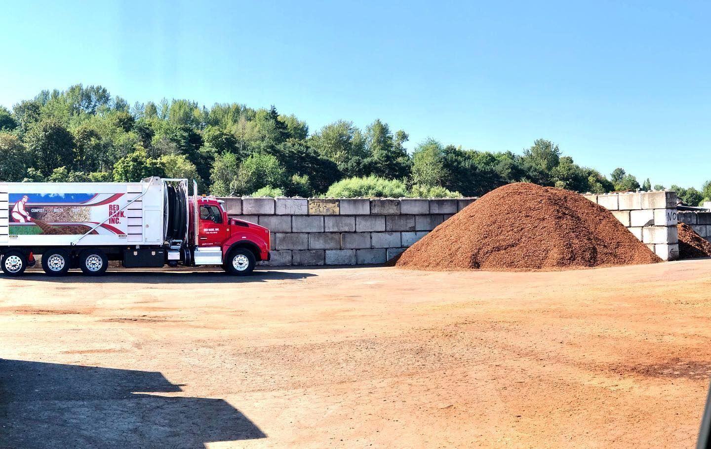 Red truck next to a pile of mulch in a gravel lot, trees in background, clear blue sky.