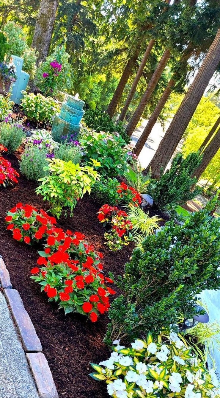 A vibrant garden bed with red and yellow flowers, green shrubs, and mulch along a sidewalk.
