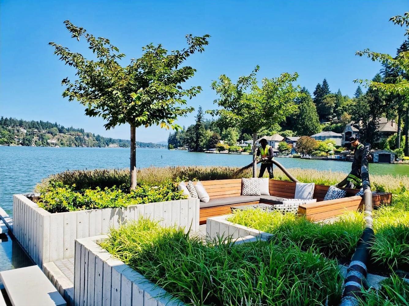 Lakeside seating area with concrete planters, wooden benches, lush greenery, and workers. Blue water and sky, trees in background.