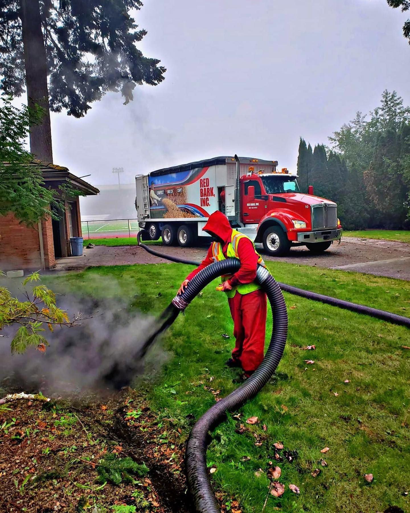 Worker in red protective suit using hose connected to a red truck, likely cleaning a septic system, outside a brick building.