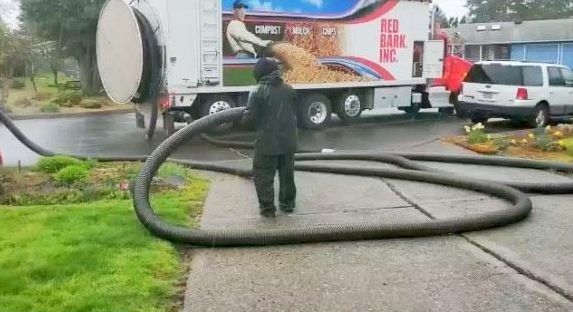 Person handling large black hose near a vacuum truck on a residential street. Wet pavement.