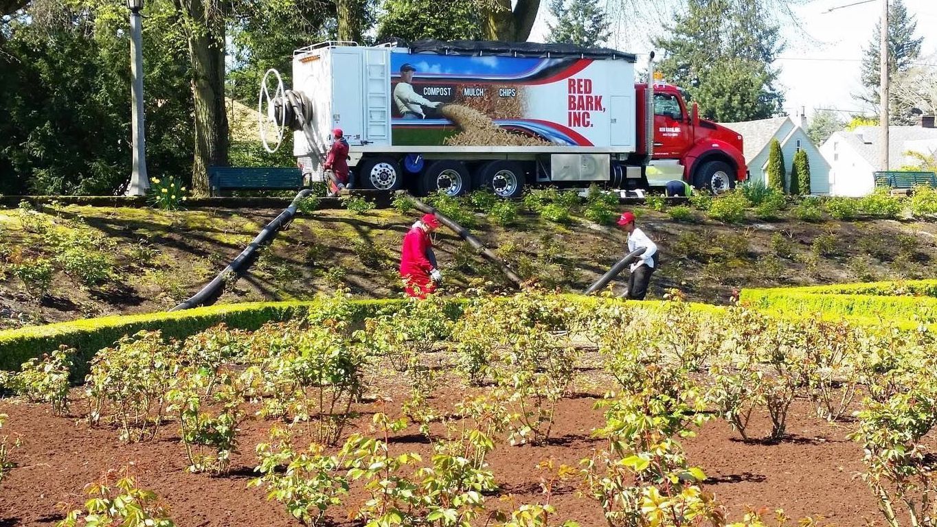 Workers in red uniforms and hard hats using equipment to clean a garden next to a large red and white truck.