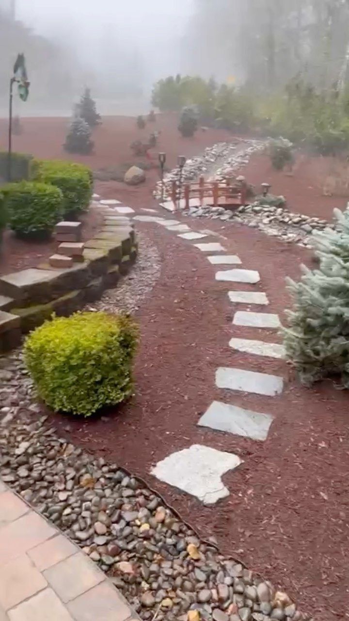 Stone pathway curves through a garden with shrubs, red mulch, and a small bridge in the fog.