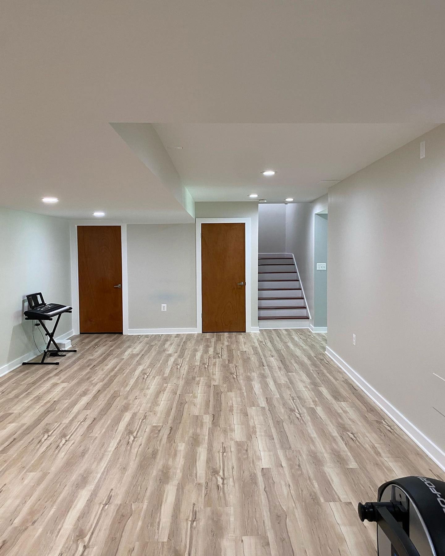 A finished basement with light wood-look flooring, neutral walls, two brown doors, and stairs leading to an upper level.