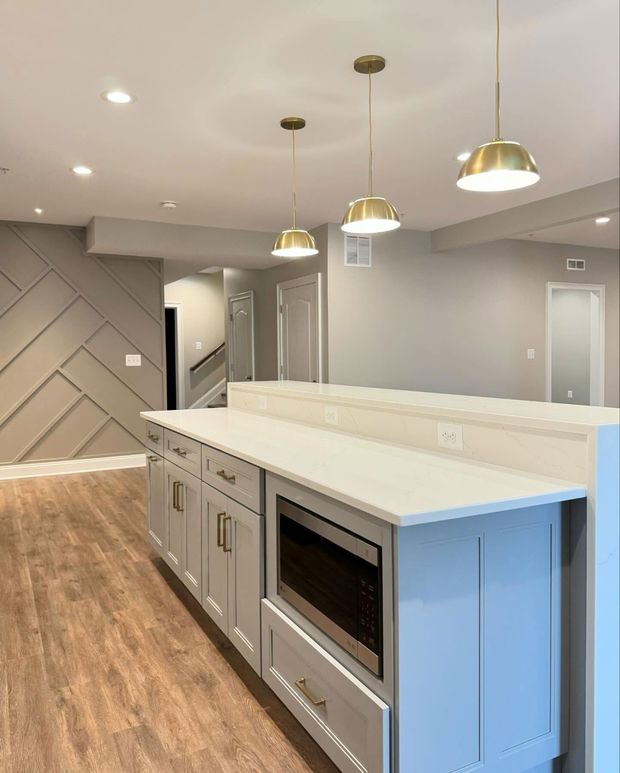 Light gray kitchen island with built-in microwave, stone countertop, gold pendant lights, and geometric wall paneling.