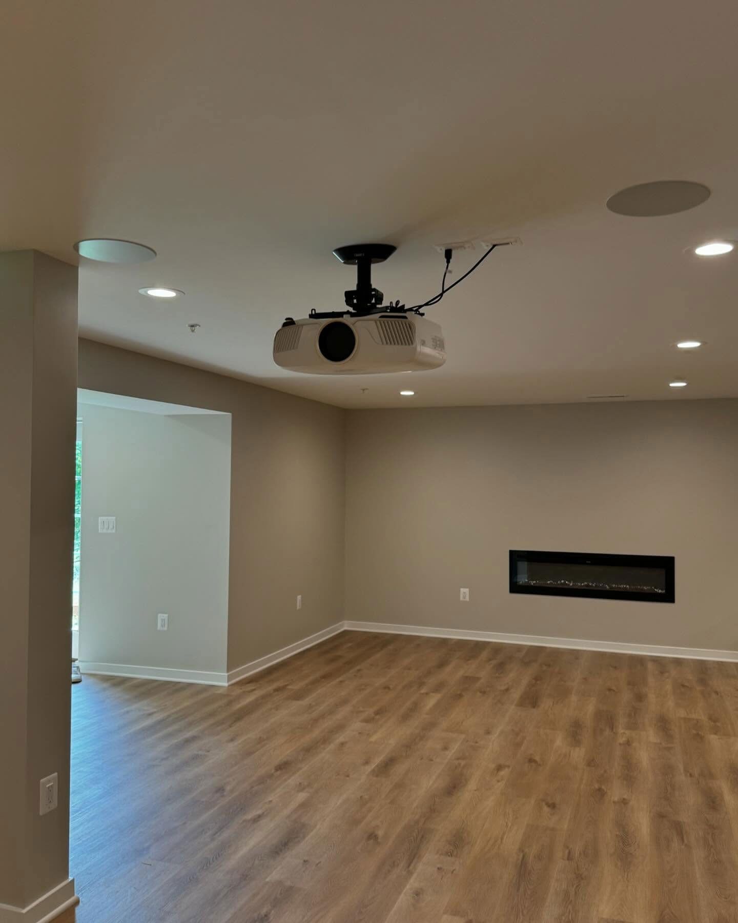 A projector mounted to the ceiling of a room with light-colored walls, wood floors, and an electric fireplace.