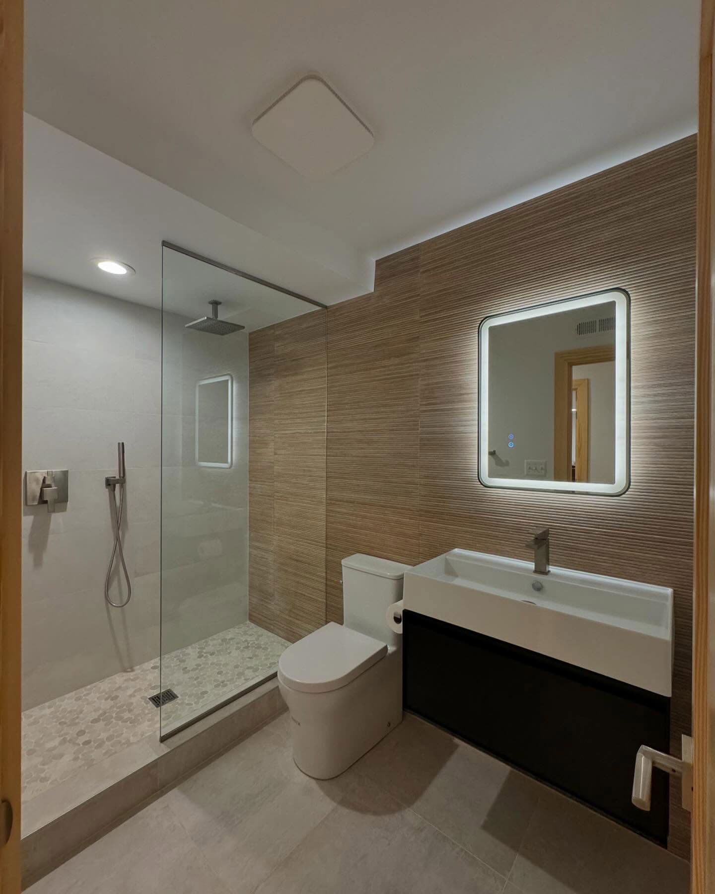 A modern bathroom featuring a walk-in shower, a toilet, and a vanity with a backlit, rounded square mirror.
