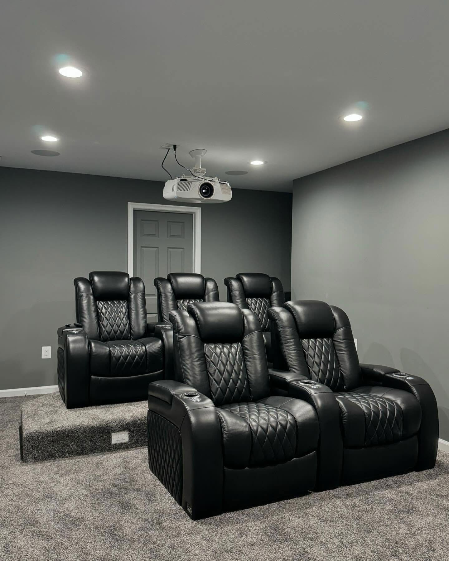 A home theater room with two rows of black leather reclining chairs on gray carpet, featuring a ceiling-mounted projector.