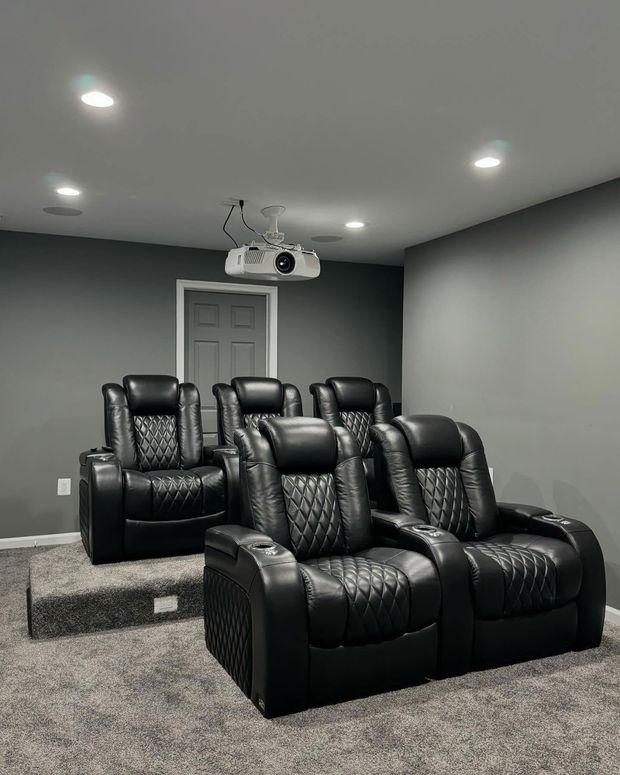 A home theater room with two rows of black leather reclining seats on grey carpet and a ceiling-mounted projector.