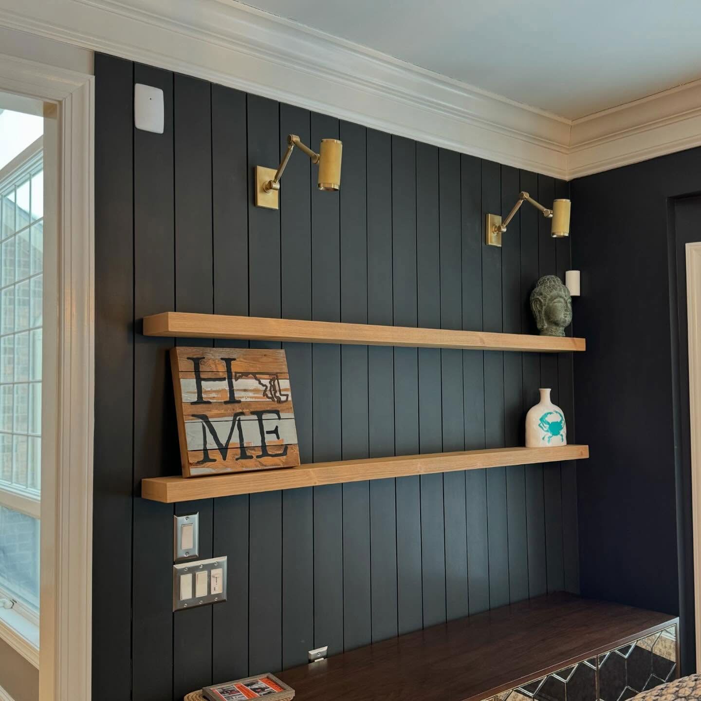 Two natural wood floating shelves mounted on a vertical, black paneled wall with gold sconces above.