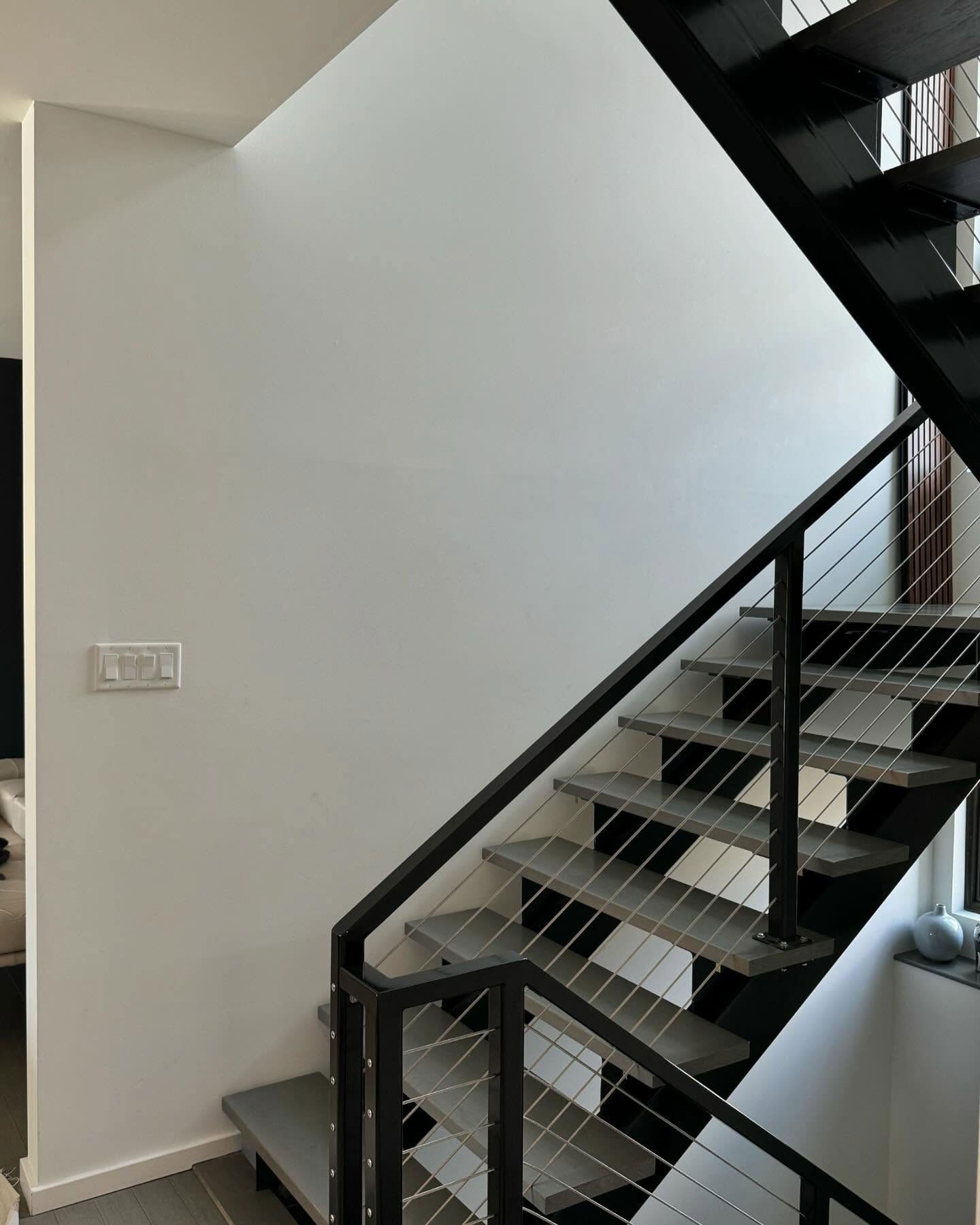 Modern indoor staircase with black metal railings, cable infill, and grey steps against a plain white wall.