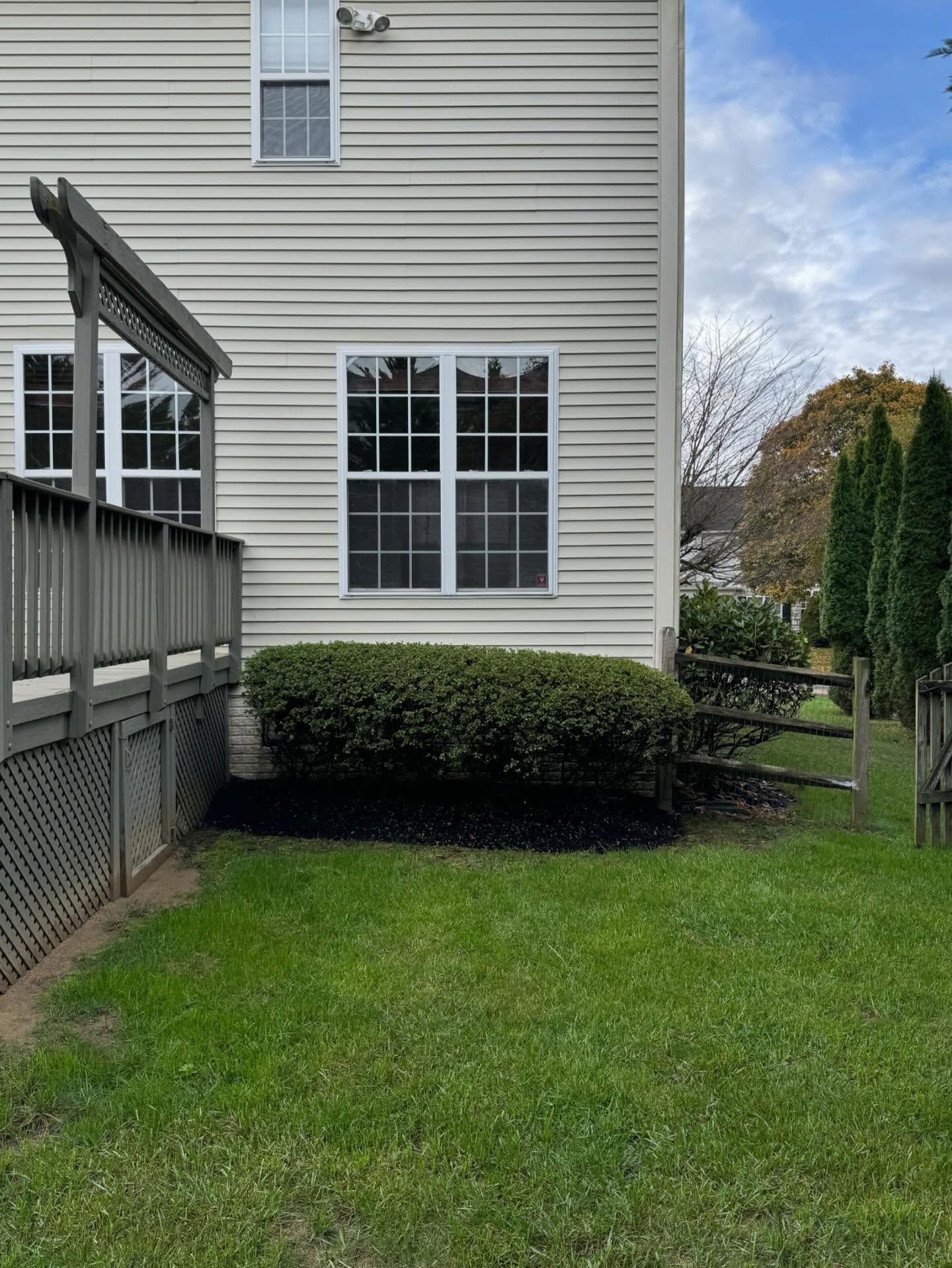 Side view of a light-colored house with a wooden deck, a hedge in a mulch bed, and a grassy yard with a wooden fence.