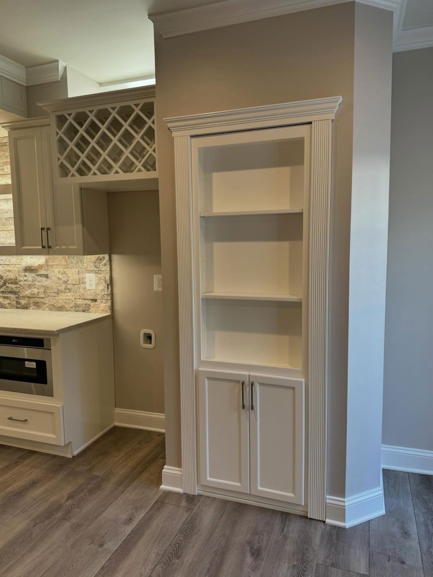 A white built-in floor-to-ceiling cabinet with shelves and lower doors, next to a kitchen counter and wine rack.