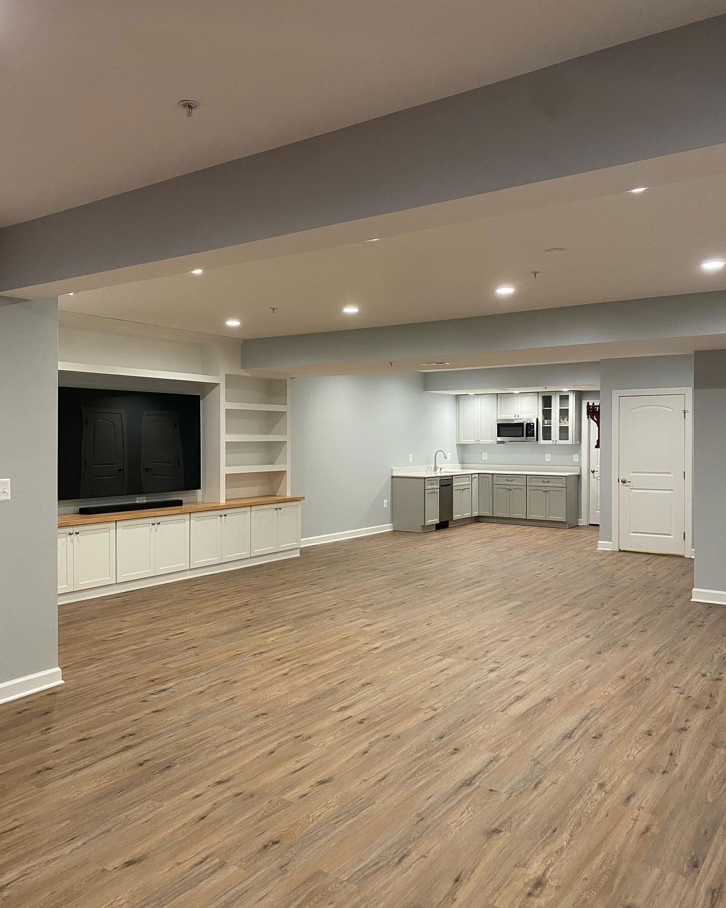 A spacious basement featuring light wood floors, a white built-in media center with a TV, and a kitchenette area.