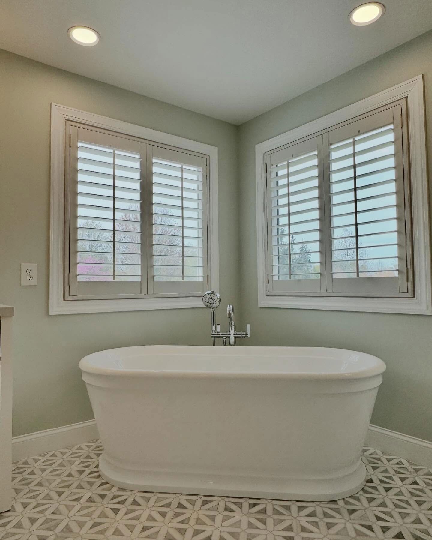 A freestanding white bathtub sits in front of two windows with white shutters in a bathroom with patterned tile floors.