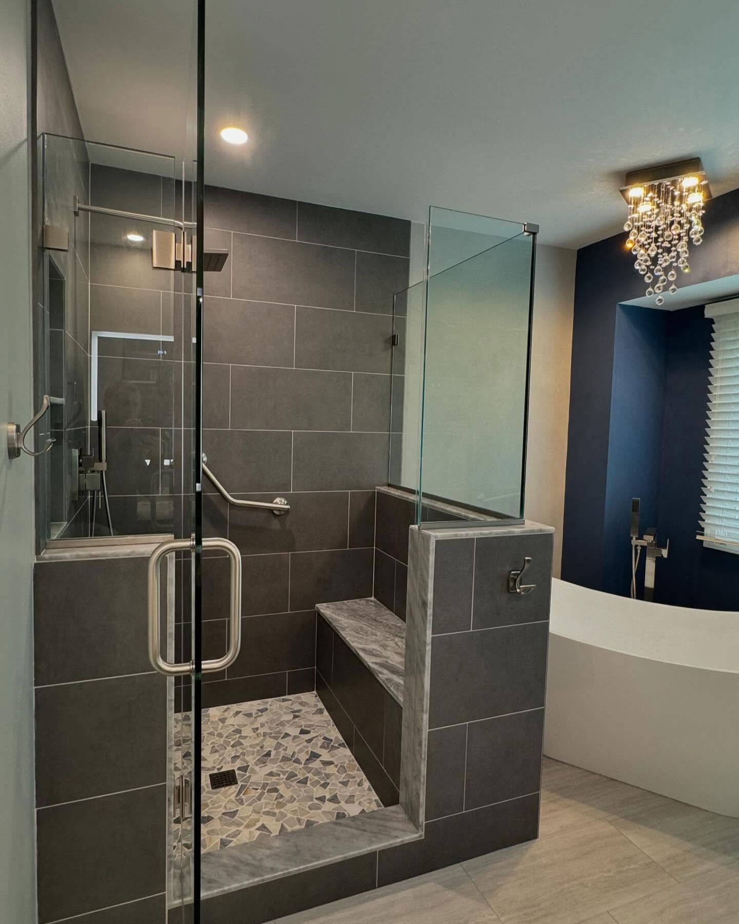 A modern bathroom featuring a tiled walk-in shower with a built-in bench, glass door, pebble floor, and a nearby bathtub.