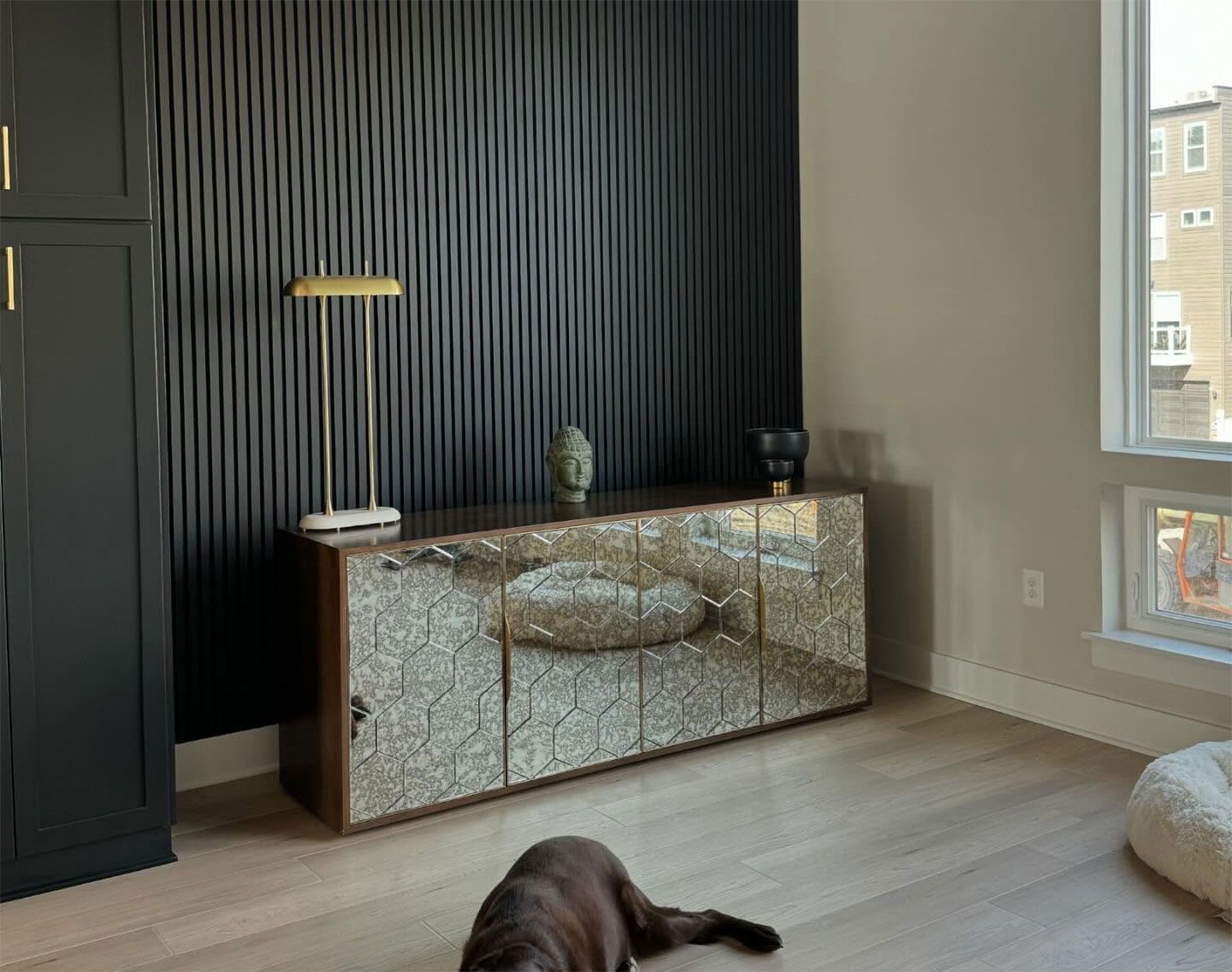 A dark-paneled accent wall behind a mirrored credenza with a brass lamp, set in a room with light wood floors and a dog.