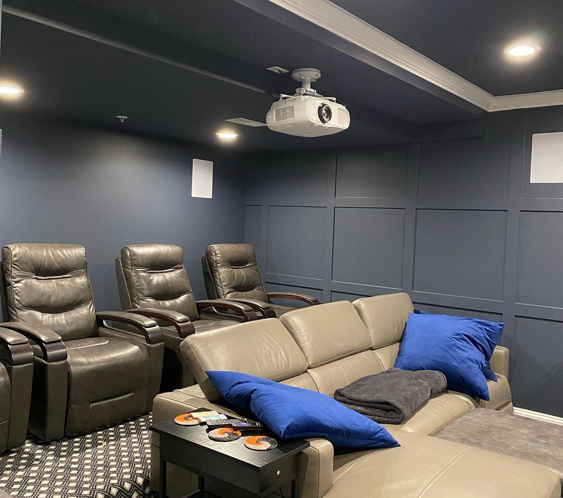 A home theater room with dark blue walls, a ceiling projector, three grey leather recliners, and a grey sectional sofa.