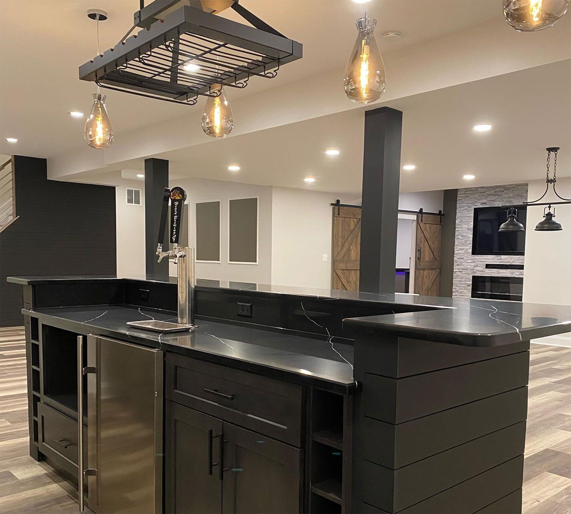 Modern basement bar with black cabinets, a beer tap, marble countertop, and pendant lighting under a ceiling rack.