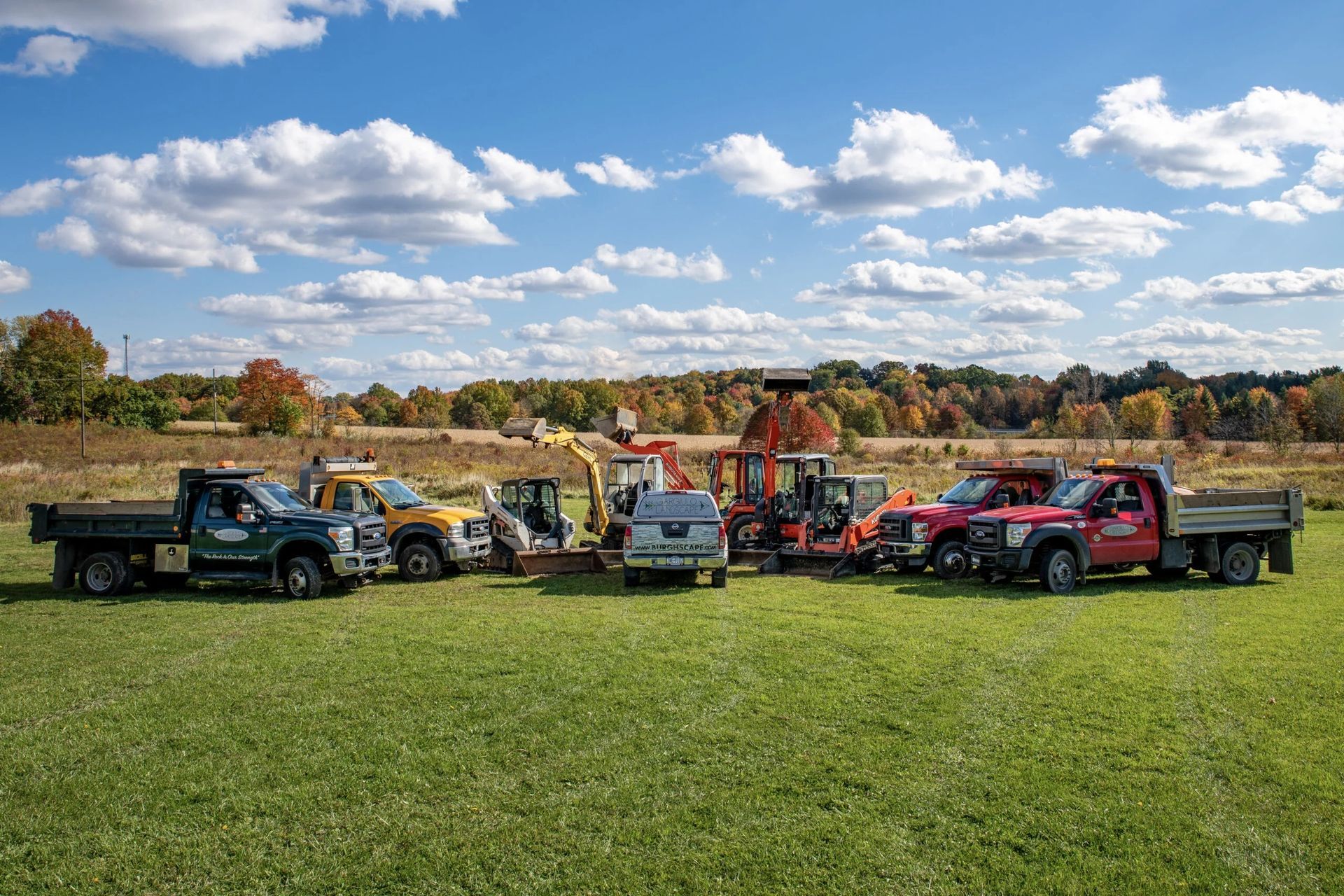 Group of construction vehicles parked on a grassy field with a backdrop of trees under a blue sky.
