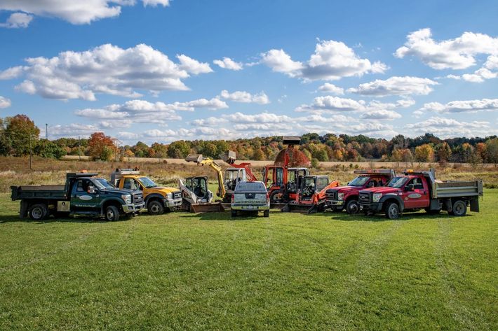 Group of construction vehicles parked on a grassy field with a backdrop of trees under a blue sky.