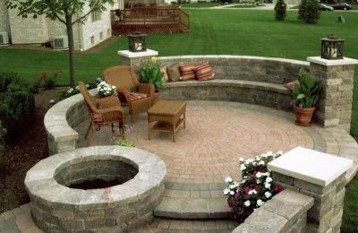 Stone patio with built-in seating, a fire pit, and potted plants. Brown and tan brickwork; green lawn backdrop.