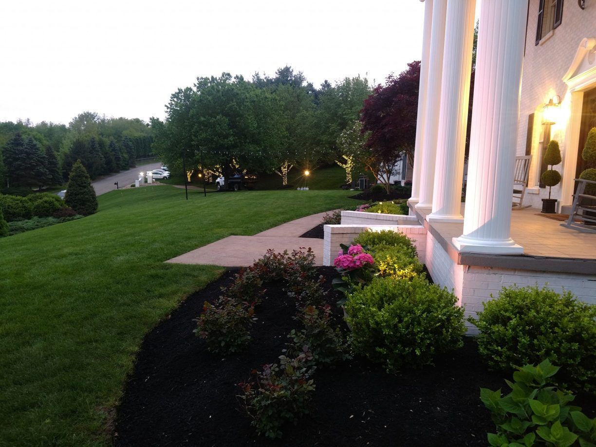 A well-manicured lawn with a white-columned house at dusk. Shrubs and flowers line the walkway.