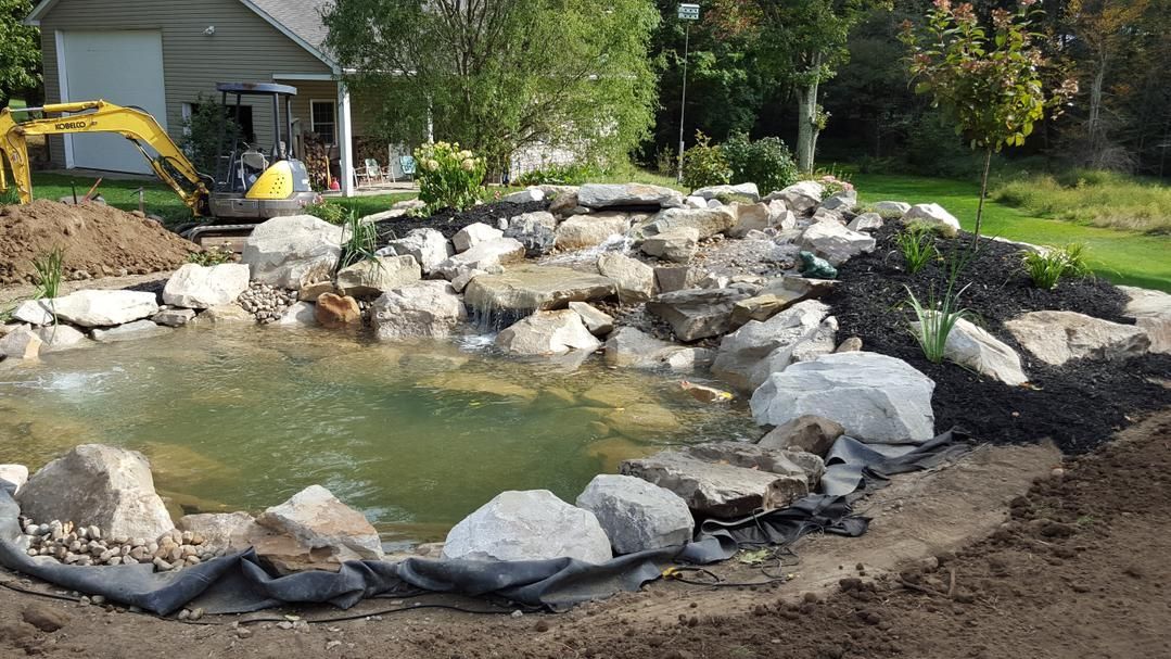 Pond with waterfall under construction, surrounded by rocks and landscaping, with an excavator in the background.