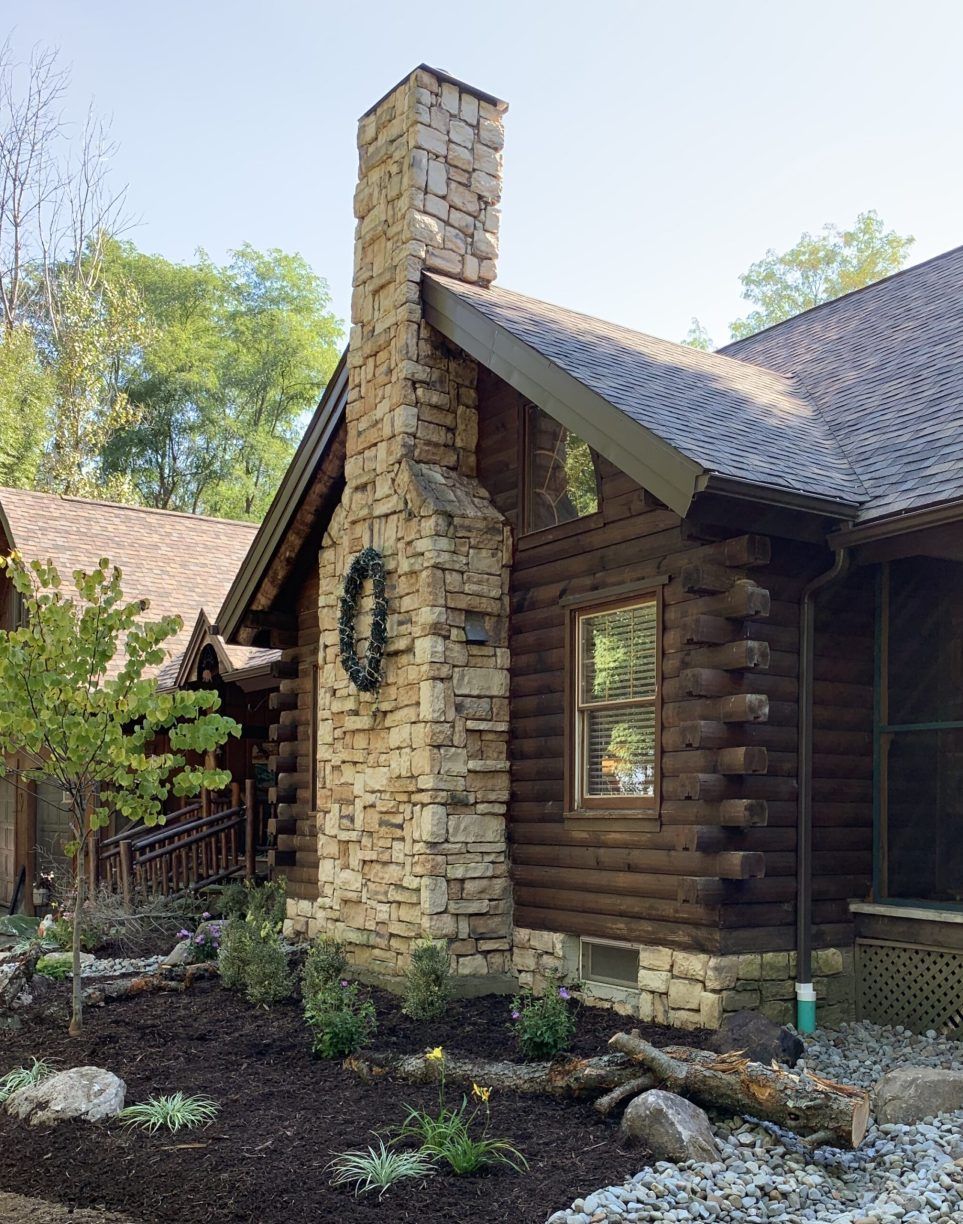 Stone chimney on a brown log cabin, surrounded by landscaping with a green wreath.