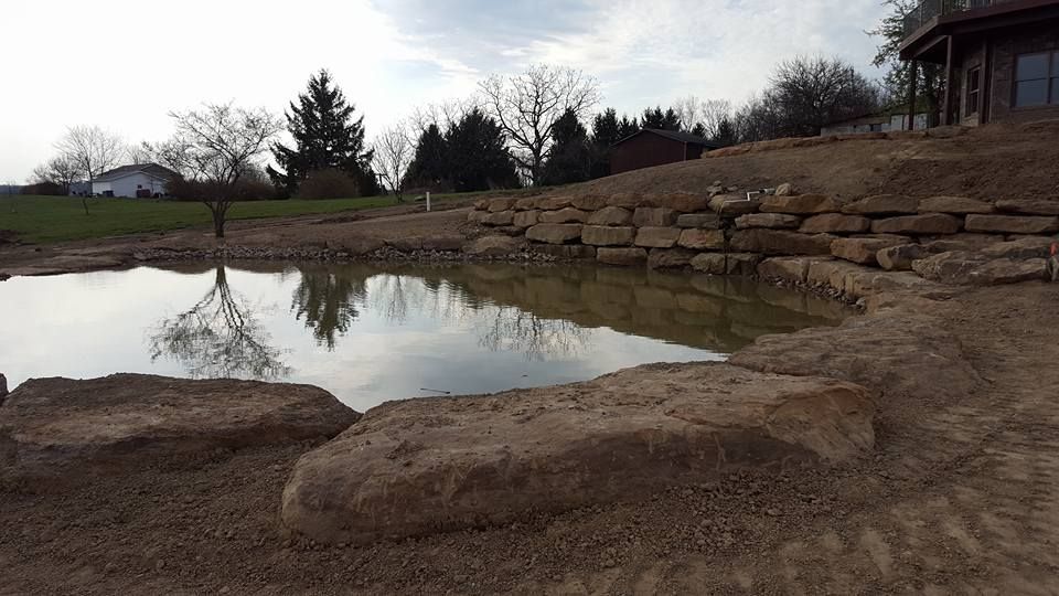 Pond surrounded by large stones and gravel, reflecting bare trees under a cloudy sky.