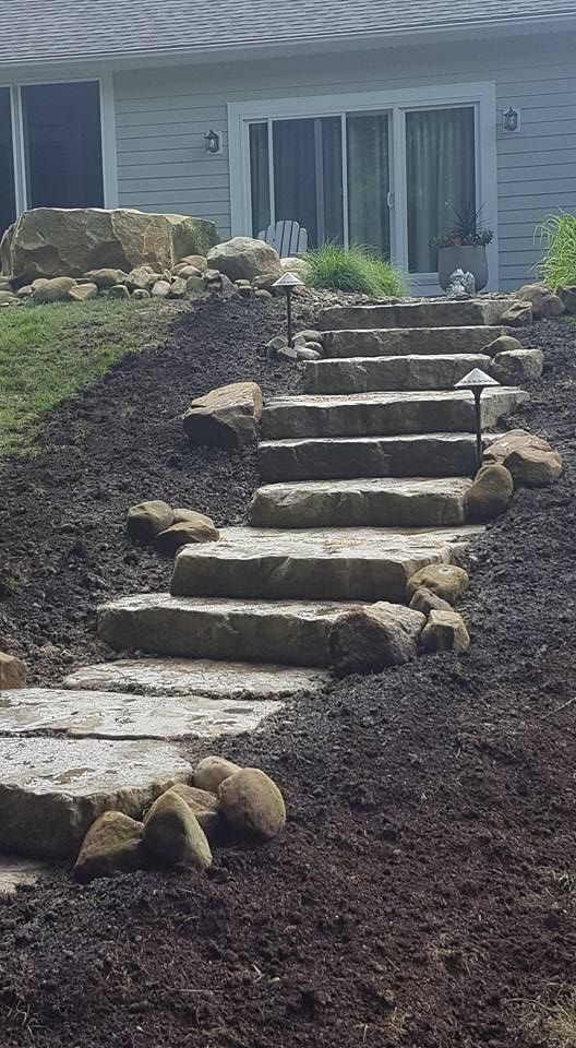 Stone steps ascend a sloped, mulched yard towards a house with sliding glass doors.