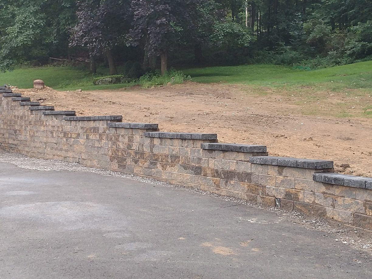 Stone retaining wall with a gray-topped edge, beside a paved surface and a grassy area with trees in the background.