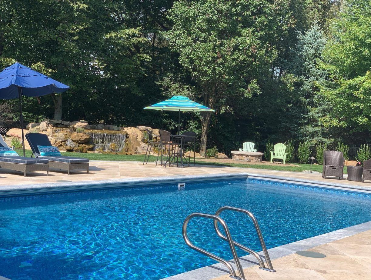 Swimming pool with blue water, lounge chairs, umbrellas, and a waterfall in a backyard setting.