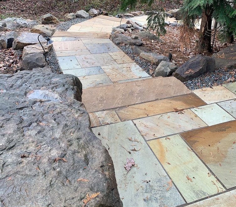 Stone pathway with steps winding through a wooded area, flanked by large rocks and greenery.