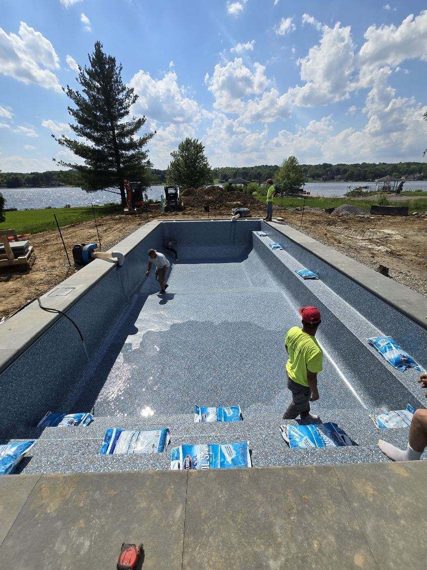 Pool construction: Workers install blue-speckled liner in rectangular pool with steps; sunny day, lake in background.