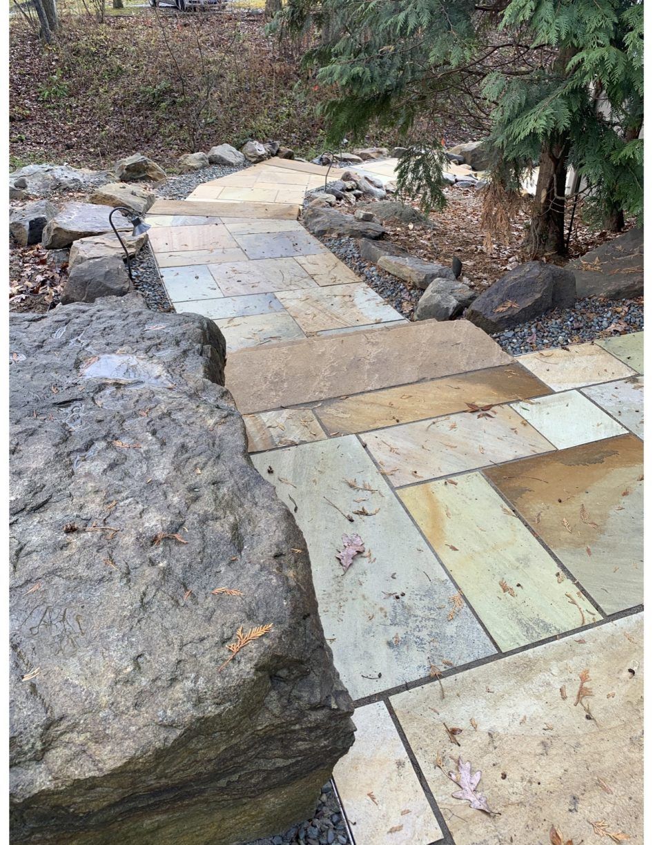 Stone pathway with stairs, edged with rocks and greenery, sloping downward.