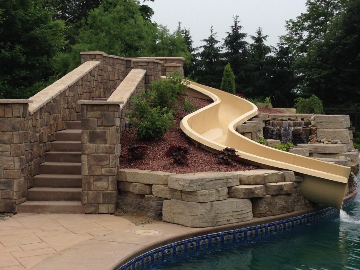 Stone staircase leading to a tan water slide built into a stone wall near a pool, surrounded by landscaping.