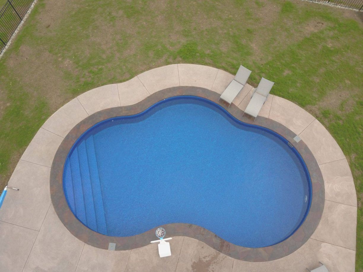 Aerial view of a blue pool with steps, surrounded by concrete and green grass; two lounge chairs are visible.