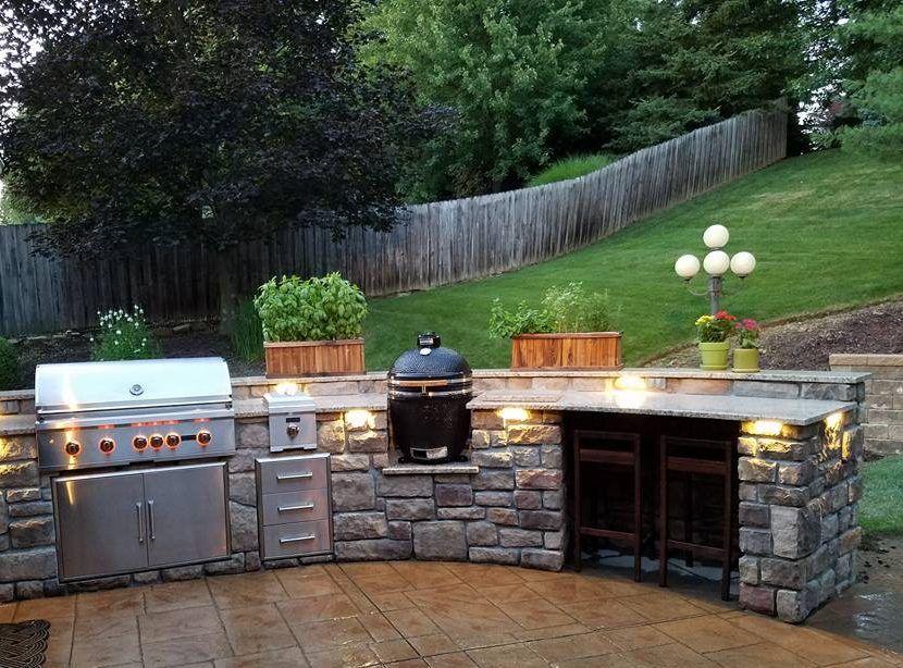 Outdoor kitchen with stainless steel grill, black egg grill, stone countertop, and seating.