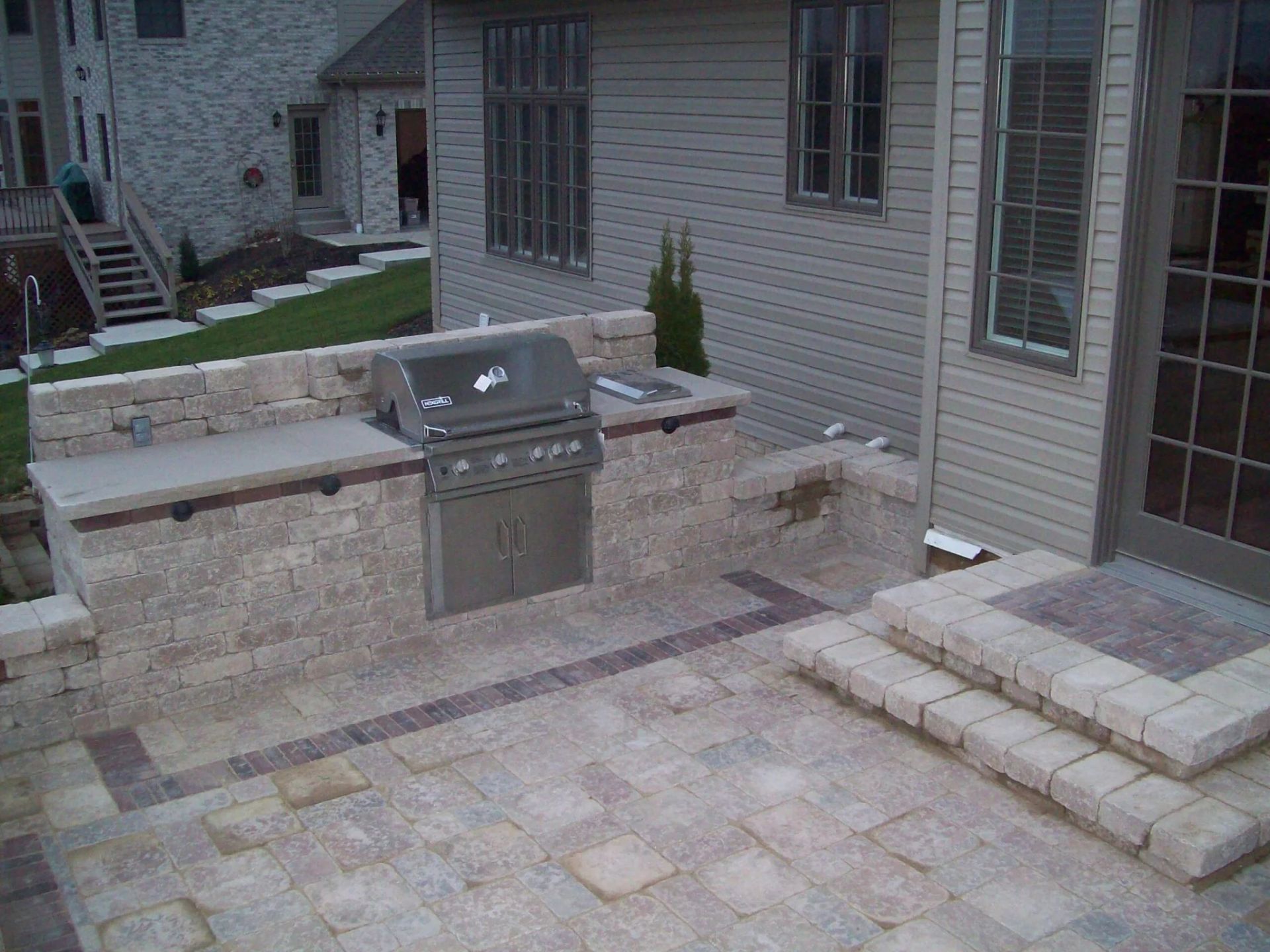 Outdoor kitchen with built-in grill on a stone patio. Steps lead to a door on a house with vinyl siding.