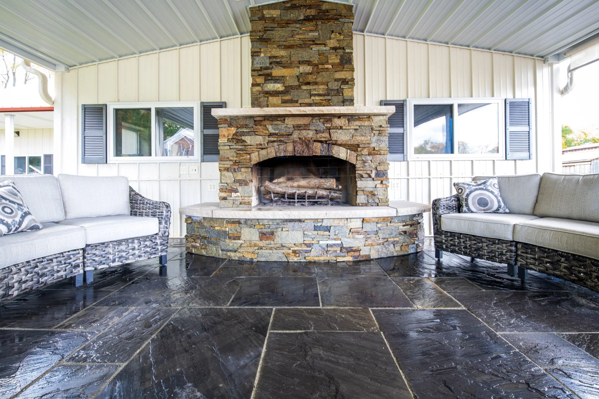 Outdoor fireplace with stone surround, flanked by two wicker couches on a dark tiled patio.