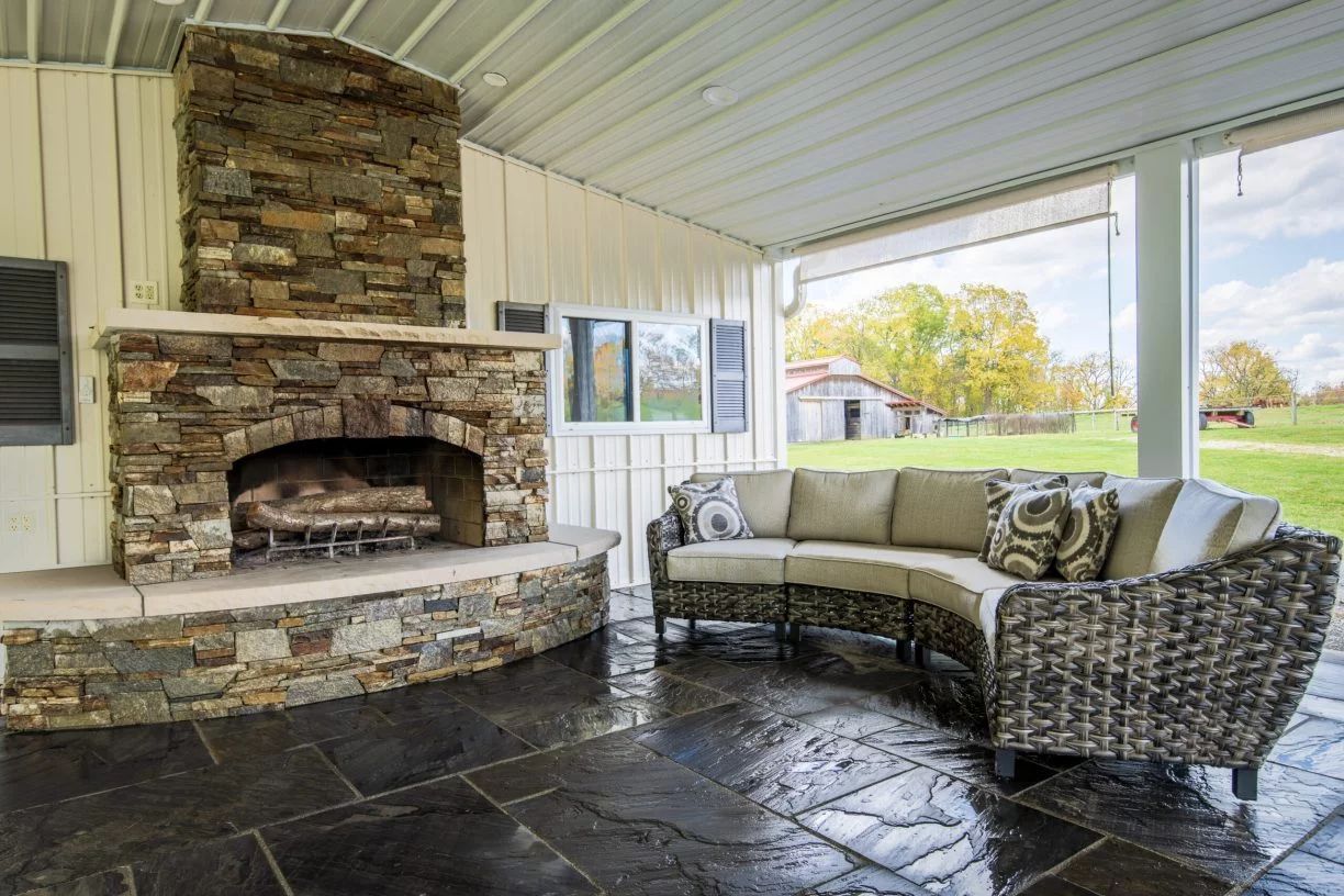 Covered outdoor patio with stone fireplace and sectional sofa overlooking a grassy field.